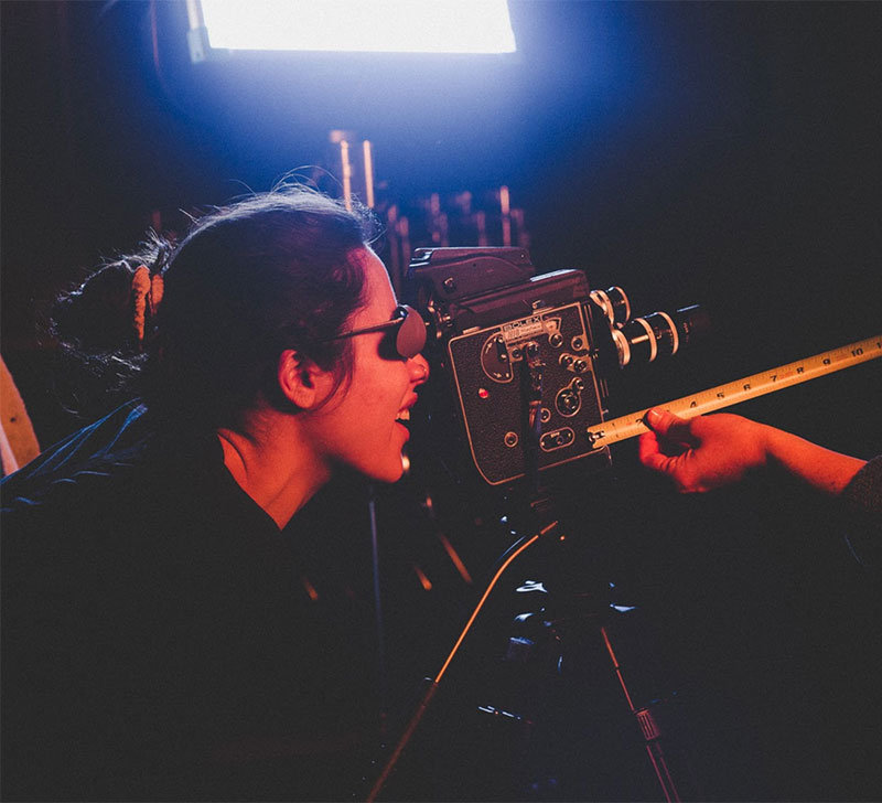 Sarah Riazati on set looking into a bolex film camera, with hands extending into frame holding a measuring tape to measure focus.