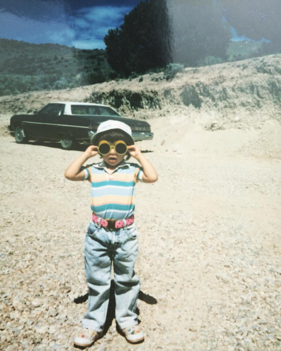 Film photo of Sarah Riazati as a toddler wearing big, hand-me-down jeans and a striped polo, holding holding yellow sunglasses up to her face against a Nevada desert landscape with an old car in the background